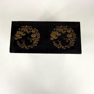 May include: A rectangular black box with two gold floral wreath designs. The box has a glossy finish and appears to be made of wood or a similar material. The floral designs are detailed and symmetrical.