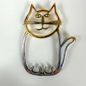 May include: A vintage cat-shaped brooch with a two-tone design. The cat's head and tail are gold-toned, while the body is silver-toned. The brooch features a simple, stylized design with a whimsical appeal. The cat has a smiling face and a curved tail.