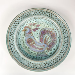 May include: A ceramic plate with a blue and white border and a stylized rooster design in shades of purple, pink, and brown.
