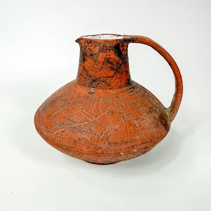 May include: A terracotta-colored ceramic pitcher with a wide, rounded base and a narrow neck. The surface features a rough, textured finish with dark, scratched markings throughout. It includes a single curved handle and a white interior rim.
