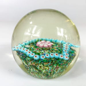 May include: A clear glass paperweight with a colorful, multi-colored, floral design inside. The design features a pink flower with blue and white flowers surrounding it.