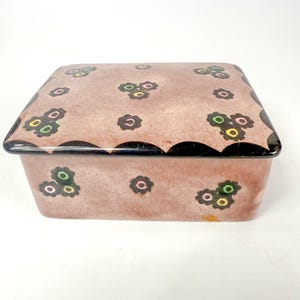 Może przedstawiać: Prostokątne ceramiczne pudełko z pokrywką. Pudełko jest w stonowanym różowym kolorze z czarną, falistą krawędzią. Pokrywka jest ozdobiona małymi kwiatowymi wzorami w kolorze zielonym, żółtym i czarnym.