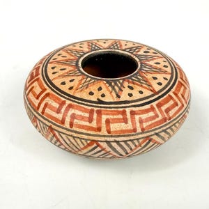 May include: A low-profile, round ceramic bowl featuring a tan base with intricate hand-painted geometric patterns in black and terracotta red. The design includes a central starburst motif, a repeating Greek key border, and linear hatching.
