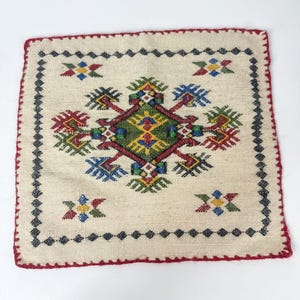 May include: Square, off-white embroidered textile with a central geometric design in green, red, yellow, and blue. The border is red, and the edges are stitched with a dark blue pattern. The textile is likely a decorative item.