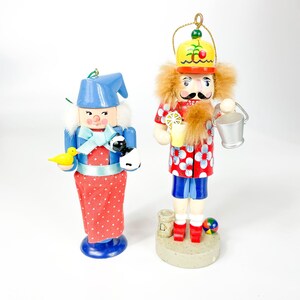 May include: Two wooden nutcracker ornaments, one is a woman wearing a blue dress with red polka dots and a blue hat, holding a yellow bird, the other is a man wearing a red and white shirt with a yellow hat and holding a bucket.