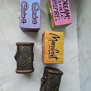 May include: An assortment of small wooden boxes with hinged lids. The boxes are painted in various colors, including blue, pink, green, and yellow. Some boxes have text that reads "Speak it Claim it", "DREAM create INSPIRE", and "Manifest THAT DREAM". Two boxes have a decorative metal design.