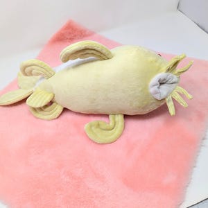 May include: A plush, yellow catfish toy with fins and whiskers, resting on a pink, fluffy surface. The toy has a light grey mouth and red eyes. The plush toy is designed to resemble a catfish.