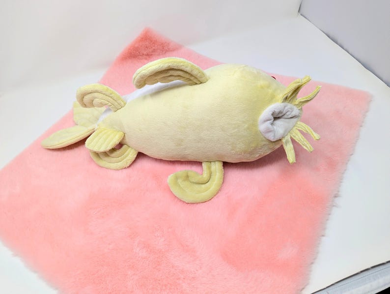May include: A plush, yellow catfish toy with fins and whiskers, resting on a pink, fluffy surface. The toy has a light grey mouth and red eyes. The plush toy is designed to resemble a catfish.