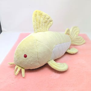 May include: A plush toy fish in shades of yellow and white, with a red eye and yellow fins. The toy is lying on a pink surface. The fish has a soft, textured appearance, suggesting it is made of fabric.