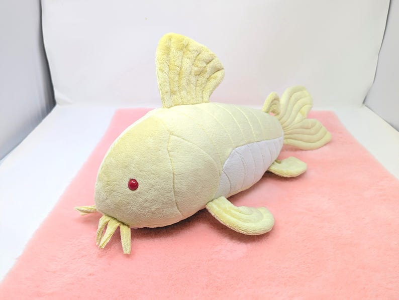 May include: A plush toy fish in shades of yellow and white, with a red eye and yellow fins. The toy is lying on a pink surface. The fish has a soft, textured appearance, suggesting it is made of fabric.