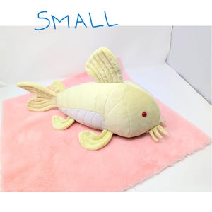 May include: A plush, yellow fish toy with red eyes and yellow fins rests on a pink, fuzzy surface. The word "SMALL" is written in blue above the fish. The toy has a light purple underbelly.