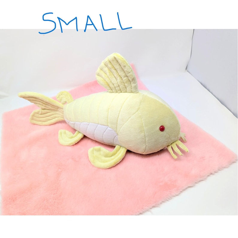 May include: A plush, yellow fish toy with red eyes and yellow fins rests on a pink, fuzzy surface. The word "SMALL" is written in blue above the fish. The toy has a light purple underbelly.