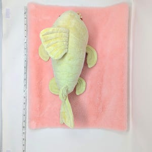 May include: A plush, light green fish toy with yellow fins and tail, resting on a pink, fluffy surface. The fish has red eyes and a small crown. The background is white.
