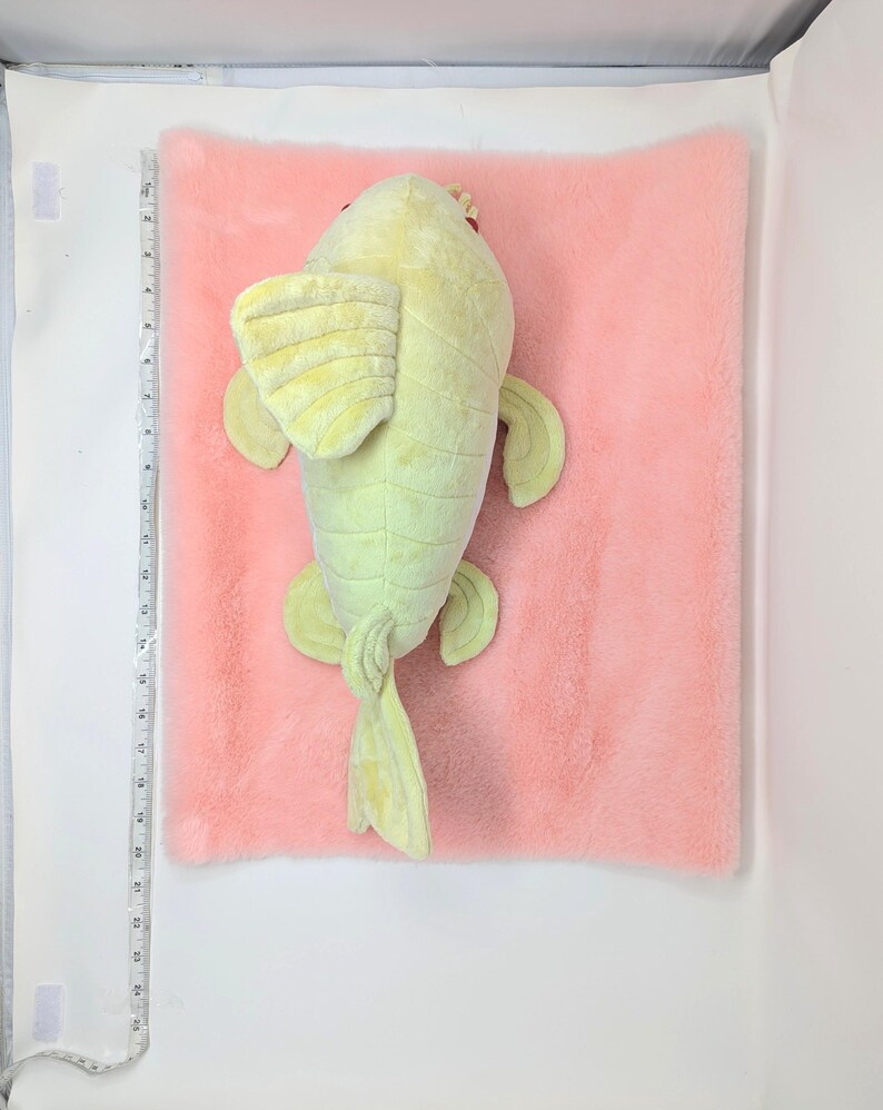 May include: A plush, light green fish toy with yellow fins and tail, resting on a pink, fluffy surface. The fish has red eyes and a small crown. The background is white.