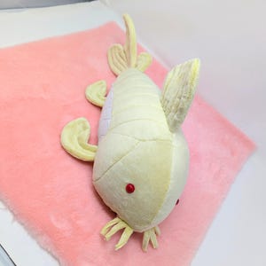 May include: A plush toy fish in shades of yellow and white with red eyes, lying on a pink, fluffy surface. The fish has fins and tail details, creating a soft, whimsical appearance. This toy is suitable for children.