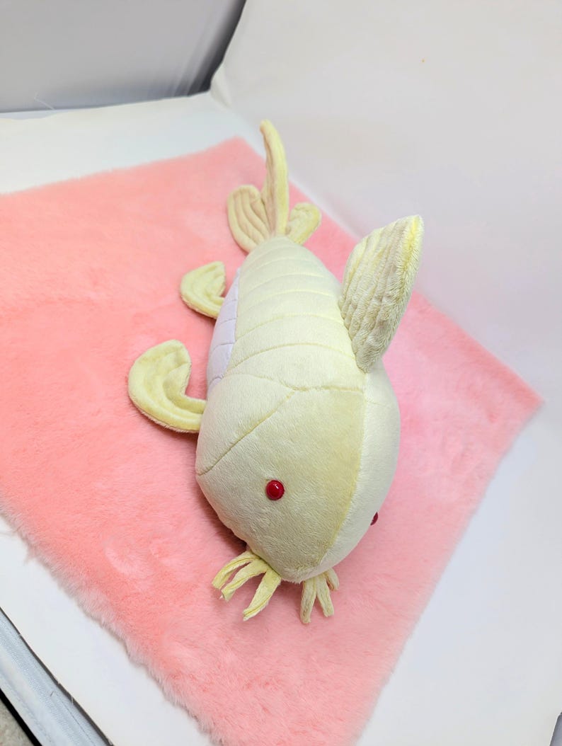 May include: A plush toy fish in shades of yellow and white with red eyes, lying on a pink, fluffy surface. The fish has fins and tail details, creating a soft, whimsical appearance. This toy is suitable for children.