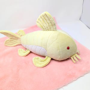 May include: A plush, yellow fish toy with red eyes and a white belly rests on a pink, fluffy surface. The fish has fins and a tail in a lighter yellow shade. The toy is designed to resemble a fish.