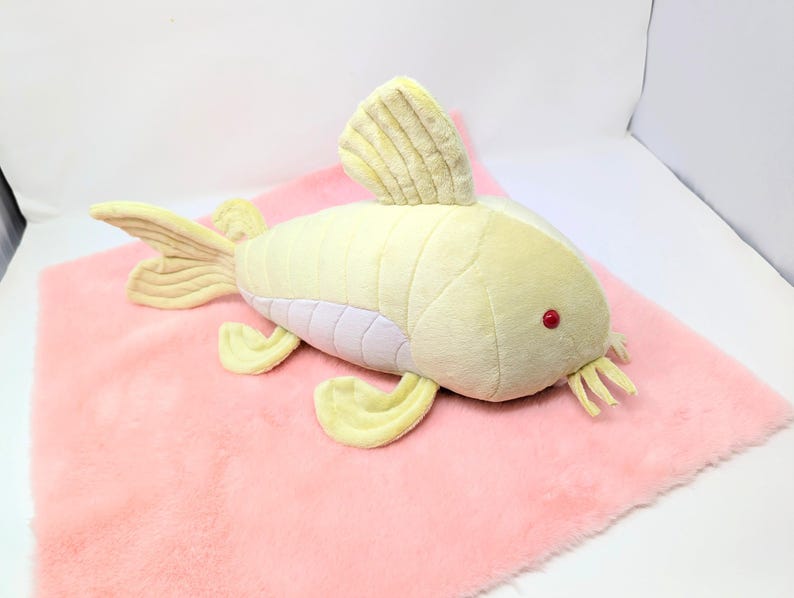 May include: A plush, yellow fish toy with red eyes and a white belly rests on a pink, fluffy surface. The fish has fins and a tail in a lighter yellow shade. The toy is designed to resemble a fish.