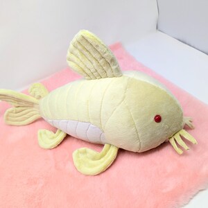 May include: A plush toy fish in shades of yellow and white, with red eyes and a white underbelly, rests on a pink, fluffy surface. The fish has fins, a tail, and whiskers. The toy is designed to resemble a catfish.