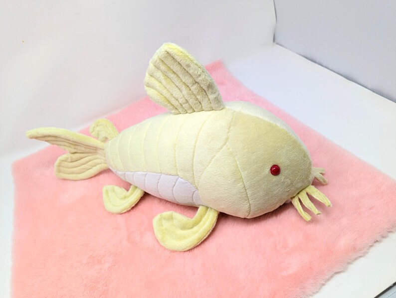 May include: A plush toy fish in shades of yellow and white, with red eyes and a white underbelly, rests on a pink, fluffy surface. The fish has fins, a tail, and whiskers. The toy is designed to resemble a catfish.