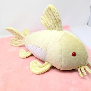 May include: A plush toy fish in shades of yellow and cream, with a red eye and cream-colored fins. The fish is lying on a pink, fuzzy surface. The toy has a soft, textured appearance.
