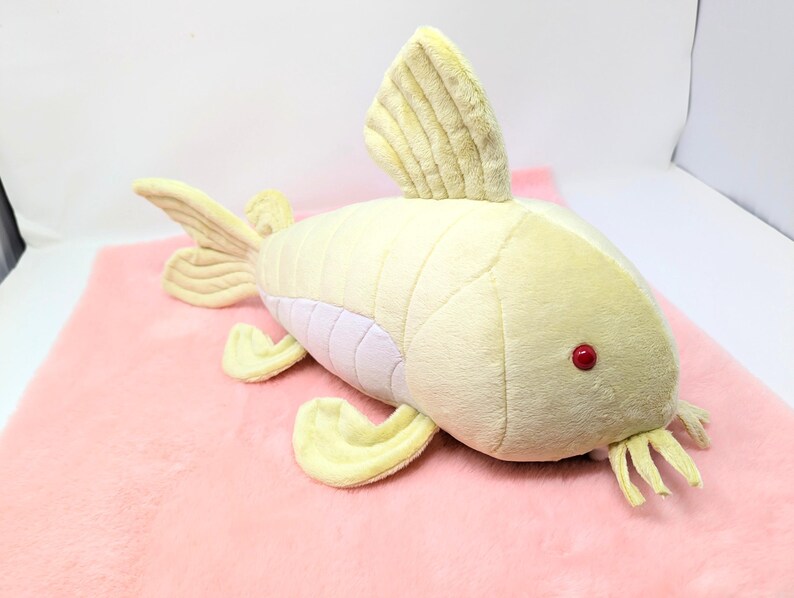 May include: A plush toy fish in shades of yellow and cream, with a red eye and cream-colored fins. The fish is lying on a pink, fuzzy surface. The toy has a soft, textured appearance.