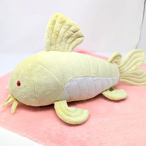 May include: A plush, yellow fish toy with a white underbelly, red eyes, and yellow whiskers. The fish has a large dorsal fin and a fan-shaped tail. It is lying on a pink surface.