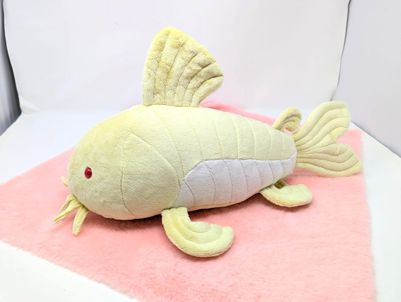 May include: A plush, yellow fish toy with a white underbelly, red eyes, and yellow whiskers. The fish has a large dorsal fin and a fan-shaped tail. It is lying on a pink surface.