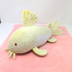 May include: A plush toy fish in shades of pale yellow and white, with red eyes and yellow fins. The toy is lying on a pink surface. The fish has a soft, textured appearance, suggesting a cuddly design.