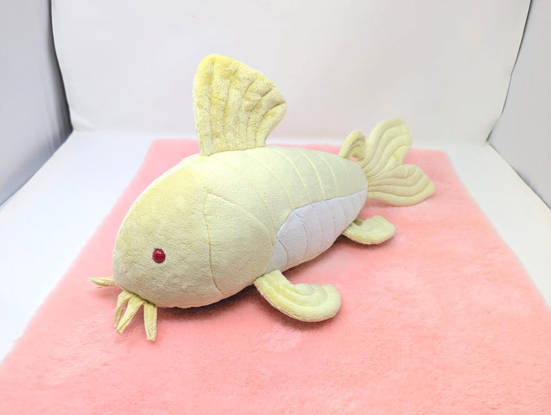 May include: A plush toy fish in shades of pale yellow and white, with red eyes and yellow fins. The toy is lying on a pink surface. The fish has a soft, textured appearance, suggesting a cuddly design.