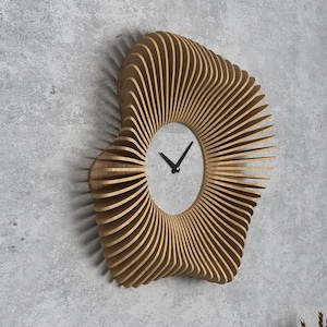 Op de afbeelding: Een houten wandklok met een uniek, abstract ontwerp. De wijzerplaat van de klok is rond en heeft zwarte wijzers op een witte achtergrond.