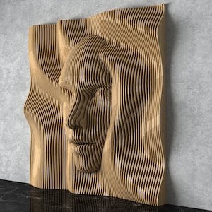 Op de afbeelding: Een houten wandsculptuur van een gezicht met een golvend, abstract ontwerp. De sculptuur is gemaakt van dunne, gelaagde stroken hout die een 3D-effect creëren.