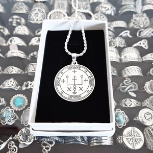 Op de afbeelding: Een zilveren ketting met een ronde hanger met een zegel met de woorden "Raphael, Adonai, Otheos, Agios" rond de rand.