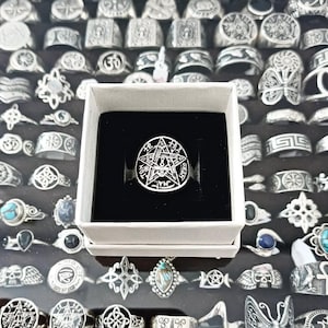 Può includere: Un anello d'argento con un design a pentagramma è esposto in una scatola bianca con interno nero. L'anello è circondato da altri anelli d'argento con vari disegni. L'anello a pentagramma presenta dettagli intricati ed è il punto focale.