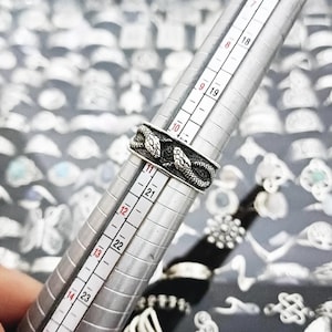 May include: A silver-toned metal ring featuring a detailed snake motif wrapped around the band. The ring is positioned on a metal ring mandrel, with the lower edge of the band aligning precisely with the size 10.75 mark on the measuring scale.