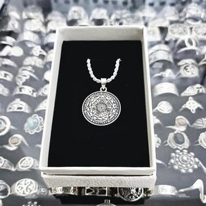 Puede incluir: Un collar de plata con un colgante redondo que presenta un diseño detallado de una estrella con formas geométricas y símbolos. El colgante está en una caja blanca con un interior negro.