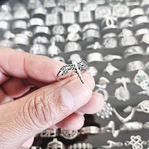 May include: A silver ring featuring a delicate dragonfly design. The ring is displayed on a finger, against a backdrop of numerous other silver rings.
