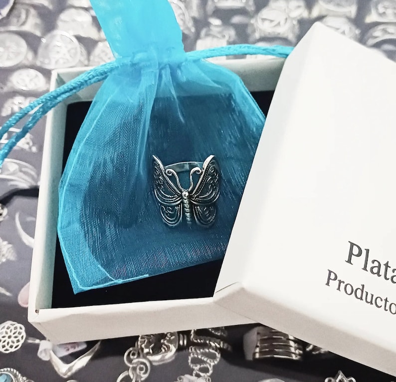 Puede incluir: Un anillo de mariposa de plata en una caja blanca con una bolsa de tul azul. La caja tiene el texto "Plata" y "Producto" impreso en ella.