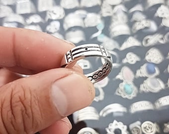 Può includere: Un anello in metallo color argento con una fascia caratterizzata da tre linee orizzontali incise, un piccolo punto centrale e accenti triangolari sui lati. La superficie interna della fascia presenta un motivo circolare ripetuto.