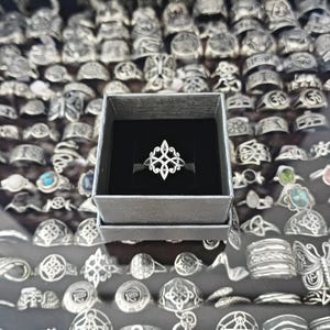 Peut inclure: Une bague en argent avec un motif ajouré, présentée dans une boîte grise avec un intérieur noir. La bague présente un motif complexe et symétrique. L'arrière-plan présente une collection de diverses bagues en argent.