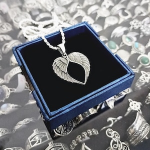 May include: A silver necklace with a heart-shaped pendant featuring angel wings. The necklace is displayed in a blue velvet box.