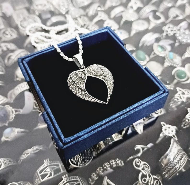 May include: A silver necklace with a heart-shaped pendant featuring angel wings. The necklace is displayed in a blue velvet box.