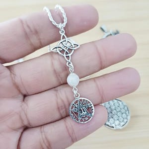 Puede incluir: Collar con colgante plateado con un nudo celta, una cuenta blanca y un dije de pentagrama. La cadena es fina. El colgante se sostiene en una mano. Una joya única.
