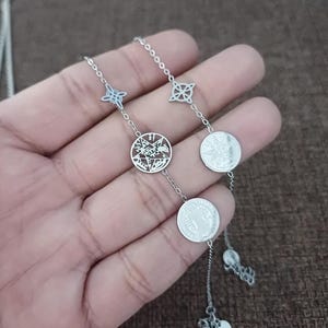 Puede incluir: Un collar plateado con una serie de colgantes. El collar incluye colgantes en forma de estrella y circulares con intrincados diseños calados. La cadena es delicada y la estética general es elegante y detallada.