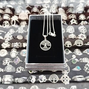Puede incluir: Un collar de tono plateado con un colgante circular que presenta un diseño recortado de una figura sentada bajo un árbol. El colgante cuelga de una cadena fina trenzada y se presenta dentro de una caja rectangular con interior de terciopelo negro.