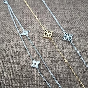May include: Close-up of three delicate bracelets. Two are silver-toned, and one is gold-toned. Each bracelet features a chain with a decorative four-petal flower charm. The bracelets are displayed on a textured brown surface.
