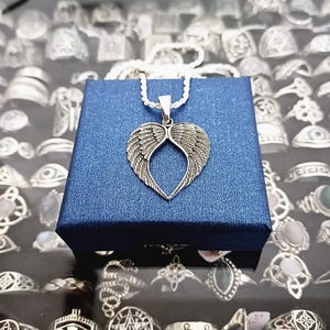 May include: Silver necklace with a heart-shaped pendant featuring detailed angel wings. The necklace is displayed on a blue velvet box.