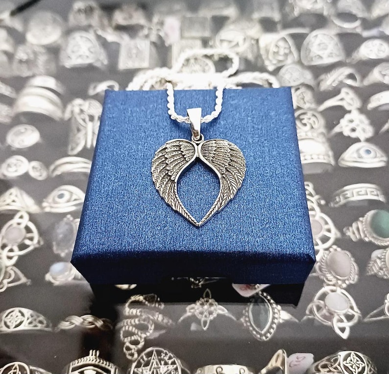 May include: Silver necklace with a heart-shaped pendant featuring detailed angel wings. The necklace is displayed on a blue velvet box.