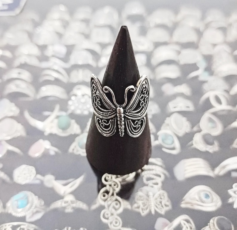 Puede incluir: Un anillo de mariposa de plata en un expositor de madera negro. El anillo est&aacute; rodeado de muchos otros anillos de plata.
