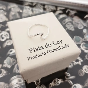 May include: A silver wave ring sits on a white box. The box has the text "Plata de Ley Producto Garantizado". The ring is shaped like a wave. Other rings are visible in the background.